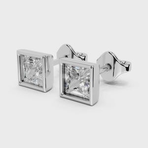 Puede incluir: Un par de pendientes de botón cuadrados con piedras preciosas transparentes. Cada pendiente presenta una piedra preciosa cuadrada engastada en un bisel plateado. Los pendientes se muestran sobre un fondo gris claro.