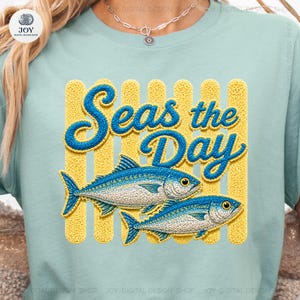 Pode incluir: Camiseta azul clara com a frase "Seas the Day" em letras azuis texturizadas, sobre dois peixes azuis e brancos. O fundo apresenta listras verticais amarelas.