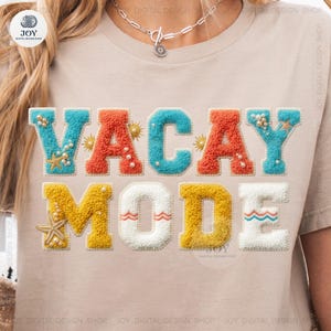Op de afbeelding: Beige T-shirt met de woorden "VACAY MODE" in kleurrijke, getextureerde letters. De letters zijn in tinten blauw, oranje, geel en wit, met decoratieve elementen zoals parels en zeesterren.