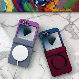 May include: Two Galaxy Z Flip5 phone cases in purple and blue. The purple case has a grey base with a white circular charging pad. The blue case has a burgundy base with a circular design. Both cases have the text "Galaxy Z Flip5".