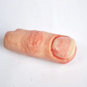 May include: A realistic prosthetic finger, flesh-toned with detailed skin texture and a pale pink nail. The finger shows subtle variations in color, with areas of pink and red, suggesting a lifelike appearance. The product is likely for special effects or medical simulation.