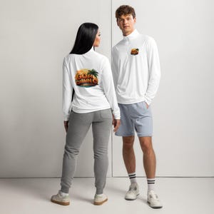 May include: Two people wearing white long-sleeve shirts and light blue shorts. The shirts have a small logo on the front and a larger graphic on the back that says "Goal Calling" with a sunset and palm tree design. The person on the left is wearing gray sweatpants.