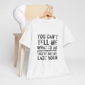 May include: White t-shirt with the text "YOU CAN'T TELL ME WHAT TO DO YOU'RE NOT MY LAST BORN" in black, distressed font. A white baseball cap rests on a wooden chair. The shirt hangs on a wooden hanger.