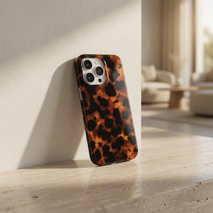 May include: A tortoiseshell-patterned phone case. The case features a glossy finish with a repeating pattern of dark brown spots on a warm, amber-colored background. The case is leaning against a white wall.