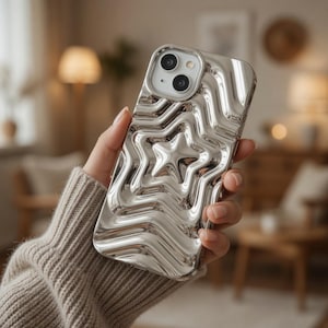 May include: A silver phone case featuring a star pattern. The case has a textured, rippled surface with a central star motif. Designed for a smartphone with dual camera lenses.