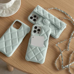 May include: A selection of pale blue quilted phone cases, including a wallet-style case with a chain strap and a case with a card pocket. The cases are displayed on a light wooden surface, showcasing a modern, stylish design.