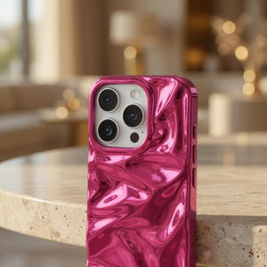 Può includere: Primo piano di una custodia per telefono rosa acceso lucida. La custodia ha una superficie strutturata e increspata e un ritaglio per la fotocamera con più obiettivi. La custodia è su una superficie chiara.