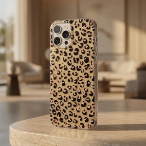 May include: A clear phone case with a leopard print design. The case features a gold background with black and pink spots. The phone case is on a light-colored stone table.