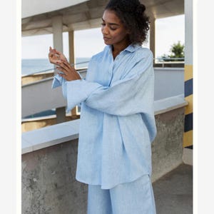 May include: A light blue long-sleeved button-down shirt with a collared neckline and wide cuffs. The garment features a relaxed, loose-fitting silhouette and a textured fabric finish, providing a simple and clean look for casual or formal wear.