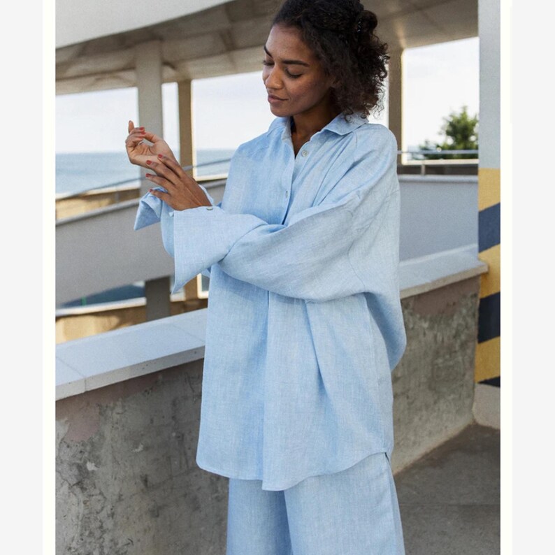 May include: A light blue long-sleeved button-down shirt with a collared neckline and wide cuffs. The garment features a relaxed, loose-fitting silhouette and a textured fabric finish, providing a simple and clean look for casual or formal wear.