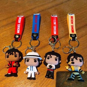 May include: Four keychains featuring miniature figures of Michael Jackson in various outfits. Each keychain has a strap with "MICHAEL JACKSON" printed on it. The figures are in red, white, black, and yellow outfits.