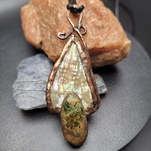 Pode incluir: Pingente artesanal com uma concha perolada envolta em cobre. O pingente tem formato de gota com uma pedra menor, em formato oval, na parte inferior. O cobre tem uma aparência rústica e envelhecida, e pende de um cordão preto.