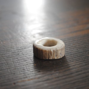 May include: A small, circular object made from a light-colored material with a hole in the center. The object has a ring shape with a textured, natural appearance, possibly made from antler or bone. The item is resting on a dark wooden surface.