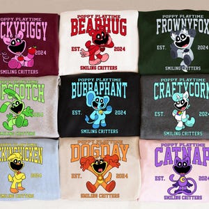 Op de afbeelding: Negen kleurrijke sweatshirts met Poppy Playtime-personages. Elk sweatshirt toont een andere karakternaam en afbeelding, samen met de tekst "EST. 2024" en "Smiling Critters". Kleuren zijn onder meer bordeauxrood, wit, groen, grijs, zwart en lichtblauw.