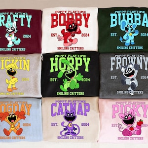 Könnte beinhalten: Neun Sweatshirts mit Charakteren aus "Poppy Playtime". Jedes Sweatshirt zeigt einen anderen Charakternamen: Crafty, Bobby, Bubba, Kickin, Hoppy, Frowny, Dogday, Catnap und Picky. Die Sweatshirts sind in verschiedenen Farben.