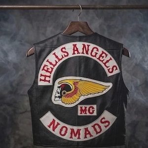 May include: Black leather vest with the words "HELLS ANGELS" and "NOMADS" in red lettering. The vest features a patch with a winged skull and the letters "MG". The vest is hanging on a wooden hanger.