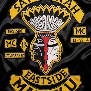 May include: A black satin jacket featuring embroidered patches with yellow and black text reading SATUDARAH, EASTSIDE, MC, 13-95-4, and EASTSIDE MALUKU. A central patch depicts a dual-faced figure in black, white, and red wearing a feathered headdress.
