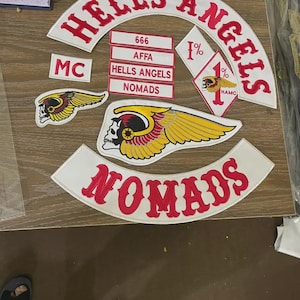 Puede incluir: Un conjunto de parches bordados con las palabras "HELLS ANGELS" en rojo sobre blanco, junto con otros parches que muestran "NOMADS", "MC", "666" y una calavera con alas. Los parches son probablemente para un club de motociclistas.