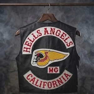 May include: Black leather vest with the words "HELLS ANGELS" in red arched text. A patch featuring a winged skull and the text "CALIFORNIA" is also visible. The vest is hanging on a wooden hanger.