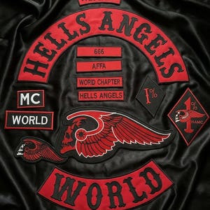 May include: A black satin jacket featuring embroidered red and black patches. The patches include the text HELLS ANGELS, NOMADS, MC, WORLD, 666, AFFA, and 1%. A large central patch depicts a winged skull, with additional winged skull and diamond-shaped 1% patches.