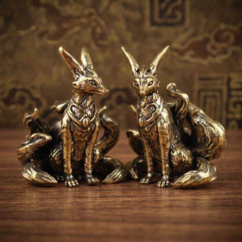 May include: Two bronze fox figurines with multiple tails, sitting upright. The foxes have detailed features, including pointed ears and ornate chest designs. The figurines are on a wooden surface, with a patterned background.