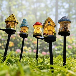 Op de afbeelding: Vijf op zonne-energie werkende tuinverlichting in de vorm van grillige huisjes en een paddenstoel. Elke lamp heeft een uniek ontwerp met levendige kleuren, waaronder geel, blauw en rood. De lampen staan op zwarte palen, met op sommige het woord "Welcome".