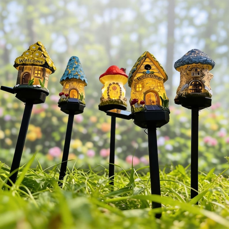 Op de afbeelding: Vijf op zonne-energie werkende tuinverlichting in de vorm van grillige huisjes en een paddenstoel. Elke lamp heeft een uniek ontwerp met levendige kleuren, waaronder geel, blauw en rood. De lampen staan op zwarte palen, met op sommige het woord "Welcome".