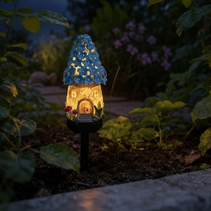 Op de afbeelding: Een zonne-aangedreven tuinpaal met een fee&euml;nhuisontwerp. Het huis heeft een beige lichaam, een blauw bloemenbedekt dak en een gloeiend interieur. Rode paddenstoelaccenten en een zwarte paal maken het ontwerp compleet.