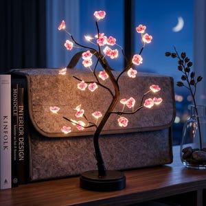 May include: A decorative LED cherry blossom tree lamp with a black base and branches. The tree features pink illuminated flowers. The lamp is on a wooden surface, with a gray bag and books in the background. The scene is lit with a soft glow.