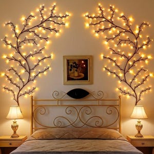 May include: A bedroom scene featuring a gold bed frame, two bedside lamps, and decorative wall lights shaped like tree branches. The branches are adorned with warm, glowing lights, creating a cozy ambiance. A framed picture hangs above the bed.