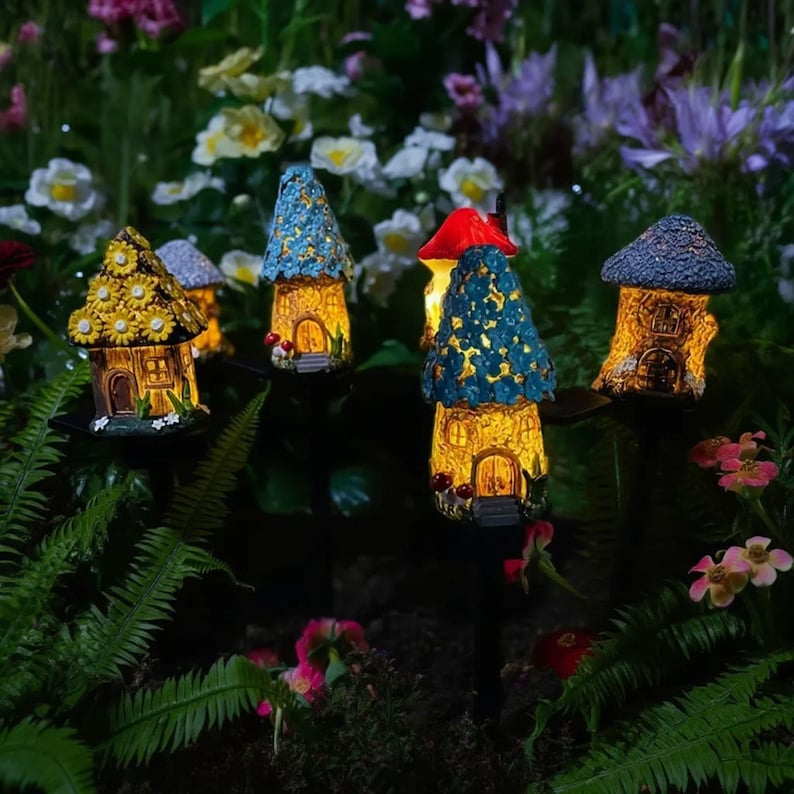 Op de afbeelding: Een verzameling verlichte miniatuurfee&euml;nhuizen met kleurrijke daken en oplichtende ramen. De huizen staan in een tuin met bloemen en groen, wat een grillige buitendecoratie cre&euml;ert. De huizen staan op palen.
