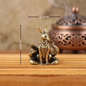 May include: A small, detailed brass fox figurine, 2.8 cm tall and 2.3 cm wide, with multiple tails and intricate design. The fox is sitting on a wooden surface, with a blurred incense burner in the background. Decorative item.