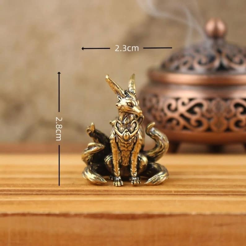 May include: A small, detailed brass fox figurine, 2.8 cm tall and 2.3 cm wide, with multiple tails and intricate design. The fox is sitting on a wooden surface, with a blurred incense burner in the background. Decorative item.