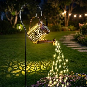 May include: A decorative outdoor solar-powered watering can with a patterned design, pouring a stream of warm white fairy lights. The metal watering can is suspended from a black metal pole, casting a patterned shadow on the grass.