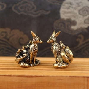 May include: Two bronze fox figurines, each with multiple tails and intricate details. The foxes are seated, facing each other, on a wooden surface. The background features a dark, patterned design. These decorative figurines are likely for home decor.