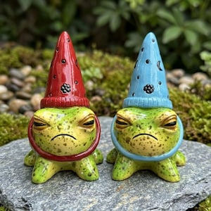 May include: Two ceramic frog figurines with grumpy expressions. One frog wears a red pointed hat with black moon and star symbols, and the other wears a light blue pointed hat with similar black celestial patterns. Both have green speckled skin.