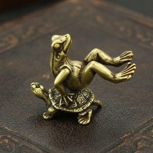 May include: A small, bronze-colored figurine of a frog riding on a turtle. The frog is in a relaxed pose, and the turtle has a detailed shell design. The figurine is likely made of metal and is a decorative item.