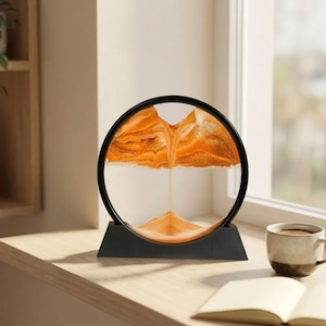 May include: A decorative sand art piece featuring orange sand within a clear glass circle, framed by a black ring and base. The sand creates a mountain-like landscape as it slowly falls. The piece sits on a wooden surface near a window.