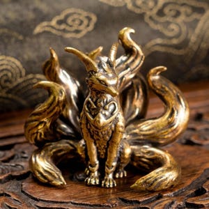 May include: A detailed brass figurine of a nine-tailed fox, a mythical creature. The fox is seated with its multiple tails fanned out. The sculpture has a metallic gold finish and sits on a wooden surface.