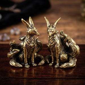 May include: Two bronze fox figurines with multiple tails, sitting upright. The foxes have detailed features, including pointed ears and intricate chest designs. The figurines are on a dark wooden surface.