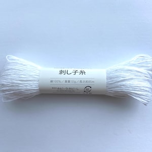 以下が含まれることがあります： 白い刺繍糸の糸巻き。日本語で「刺し子糸」と書かれたラベルが付いています。ラベルには、糸が綿100％で、重さ10g、長さ約85mと記載されています。白い背景に糸巻きが置かれています。