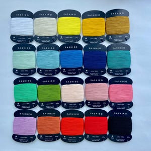 May include: Twenty spools of Sashiko thread arranged in a grid, each wrapped around a black cardboard card. The collection features a diverse spectrum of colors including white, yellow, blue, green, pink, orange, red, and black, all labeled 100% cotton.
