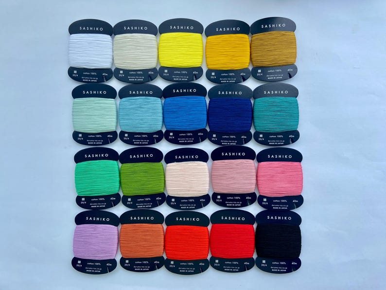 May include: Twenty spools of Sashiko thread arranged in a grid, each wrapped around a black cardboard card. The collection features a diverse spectrum of colors including white, yellow, blue, green, pink, orange, red, and black, all labeled 100% cotton.