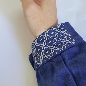 K&ouml;nnte beinhalten: Ein dunkelblaues lang&auml;rmeliges Kleidungsst&uuml;ck mit einer strukturierten Manschette, die mit aufwendiger wei&szlig;er geometrischer Stickerei verziert ist. Die Stiche bilden ein wiederkehrendes Muster aus ineinandergreifenden Kreisen und kleinen Kreuzen.