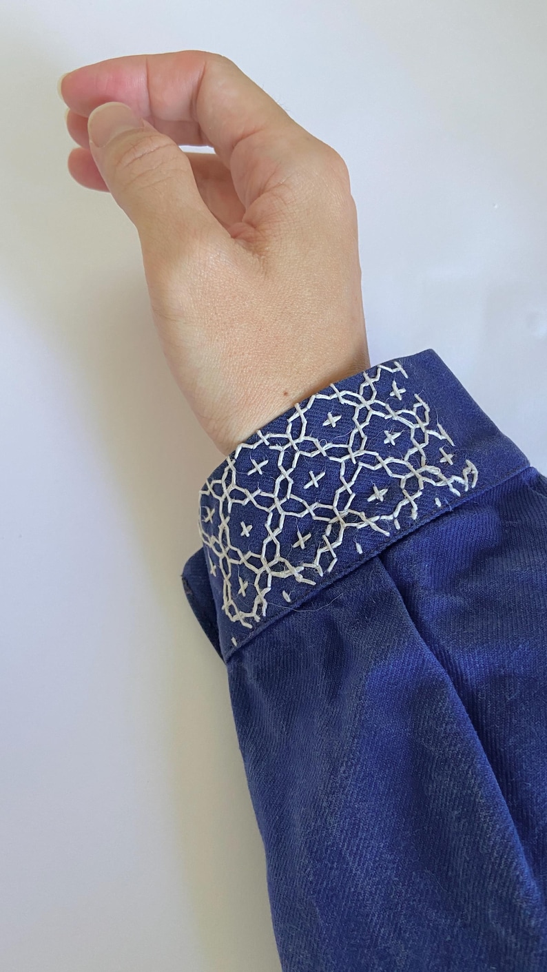 K&ouml;nnte beinhalten: Ein dunkelblaues lang&auml;rmeliges Kleidungsst&uuml;ck mit einer strukturierten Manschette, die mit aufwendiger wei&szlig;er geometrischer Stickerei verziert ist. Die Stiche bilden ein wiederkehrendes Muster aus ineinandergreifenden Kreisen und kleinen Kreuzen.