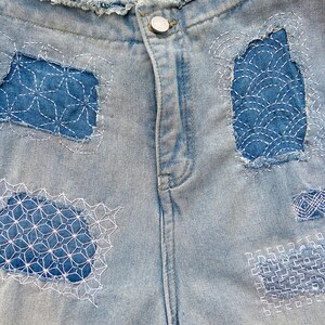 K&ouml;nnte beinhalten: Hellblaue Jeansshorts mit ausgefranstem Bund und einem silberfarbenen Metallknopf. Der Stoff ist mit vier verschiedenen Flicken aus dunklerem blauem Denim verziert, die jeweils mit aufwendigen wei&szlig;en geometrischen Sashiko-Stickereien versehen sind.