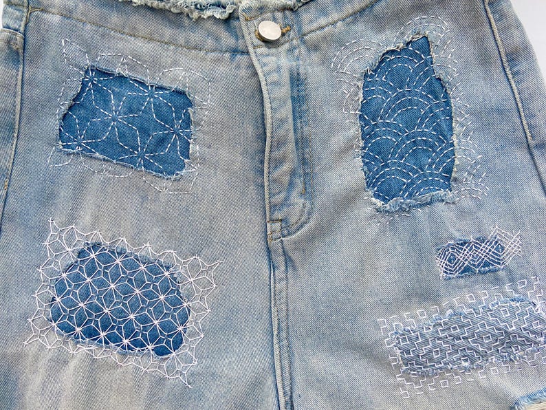 K&ouml;nnte beinhalten: Hellblaue Jeansshorts mit ausgefranstem Bund und einem silberfarbenen Metallknopf. Der Stoff ist mit vier verschiedenen Flicken aus dunklerem blauem Denim verziert, die jeweils mit aufwendigen wei&szlig;en geometrischen Sashiko-Stickereien versehen sind.