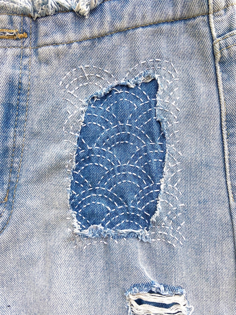 May include: Light blue denim shorts featuring a distressed rectangular patch with a dark blue denim insert. The insert is decorated with white sashiko-style stitching in a repeating fan pattern. The shorts have a frayed hem and additional distressing.