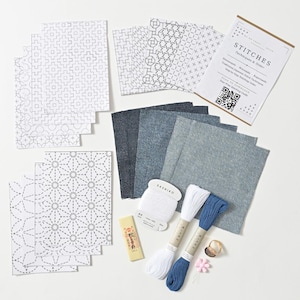 Puede incluir: Un kit de bordado Sashiko que contiene varios cuadrados de tela con patrones geométricos impresos, varias muestras de tela color mezclilla, dos madejas de hilo, un carrete de hilo blanco, un marcador de tiza amarillo, un dedal y una tarjeta de instrucciones con un código QR etiquetado como STITCHES Techniques & Order.