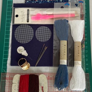May include: A selection of sewing tools and materials, including thread spools, needles, fabric pieces, and a pink marking pen. The items are arranged on a green cutting mat, displaying a range of colours and textures suitable for craft projects.